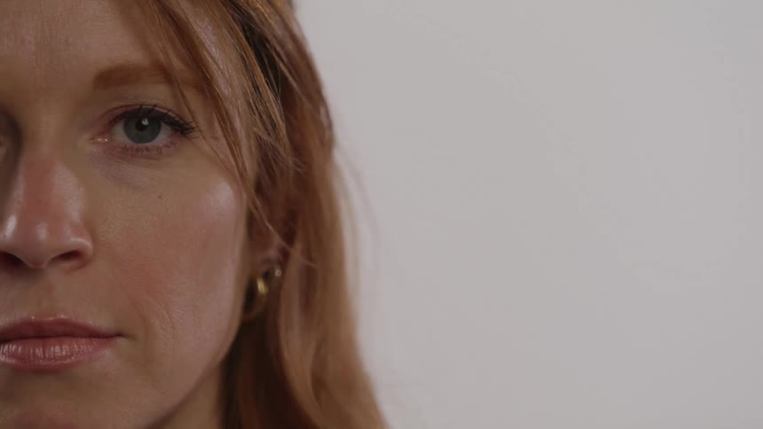 A closeup shot focuses on the left side of a womans face showing her with auburn hair and a small hoop earring her eye opens quickly changing her expression from one of quiet contemplation to one of direct engagement with the camera