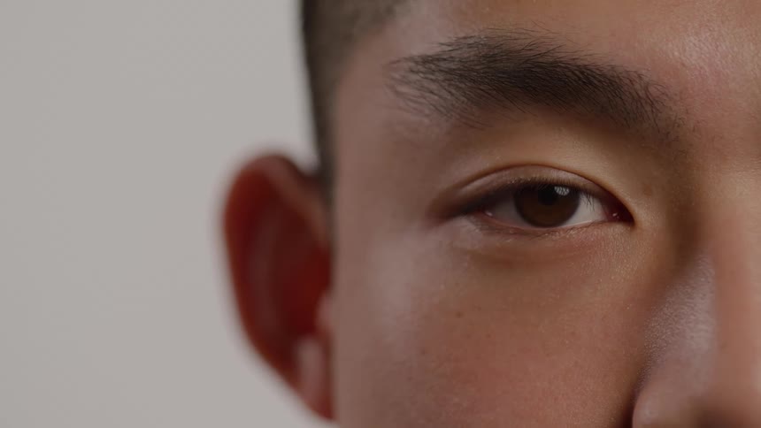 This is an extreme closeup shot of a persons face focused on their right eye and ear against a plain background the eye slowly opens from a closed state to a steady thoughtful gaze toward the camera emphasizing the detail of the brown iris and dark