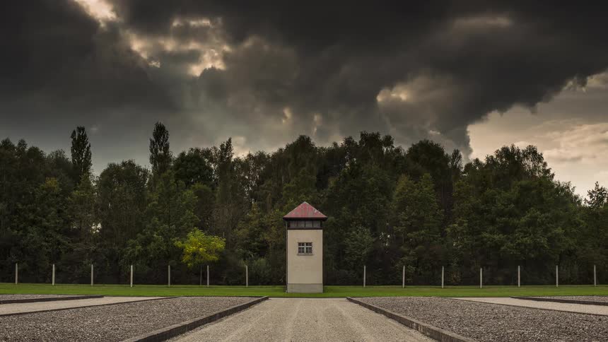 Dark stormy clouds roll rapidly across the sky in a dramatic timelapse passing over a solitary guard tower that stands between a dense forest and wide gravel paths