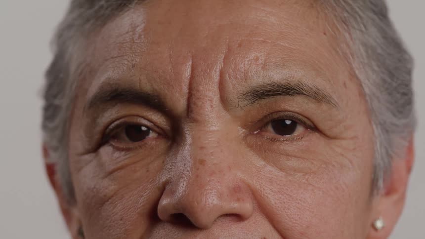 The video is a tight closeup of an older persons face focusing on their eyes and forehead against a plain background the person slowly opens their eyes revealing a direct gaze and the deep lines on their forehead