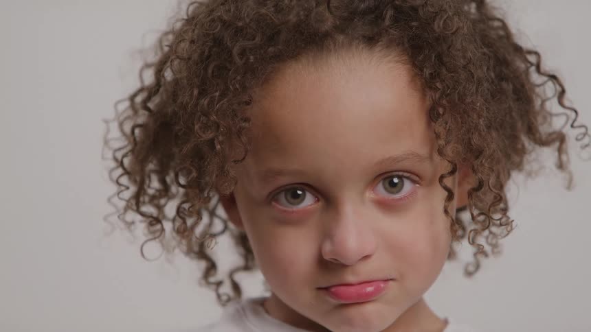 A young child with curly hair puckering their lips and looking directly ahead with a serious expression