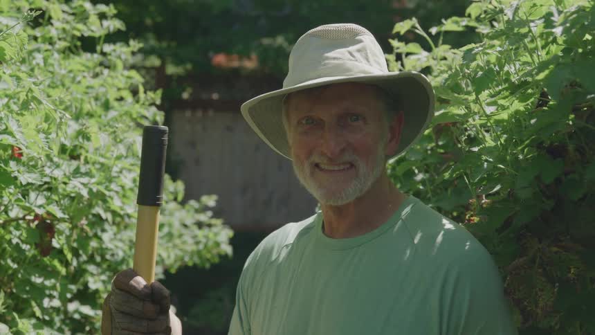 A smiling middleaged man wearing a lightcolored sun hat and a tshirt stands in a sunny garden holding a garden tool with a black and yellow handle he looks directly at the camera while surrounded by lush green foliage