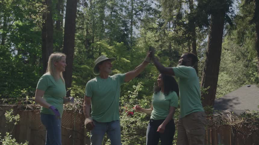 A group of four adults all wearing light green shirts and work gloves are celebrating their gardening efforts by giving each other high fives they are standing outdoors in a lush sunny yard surrounded by tall trees