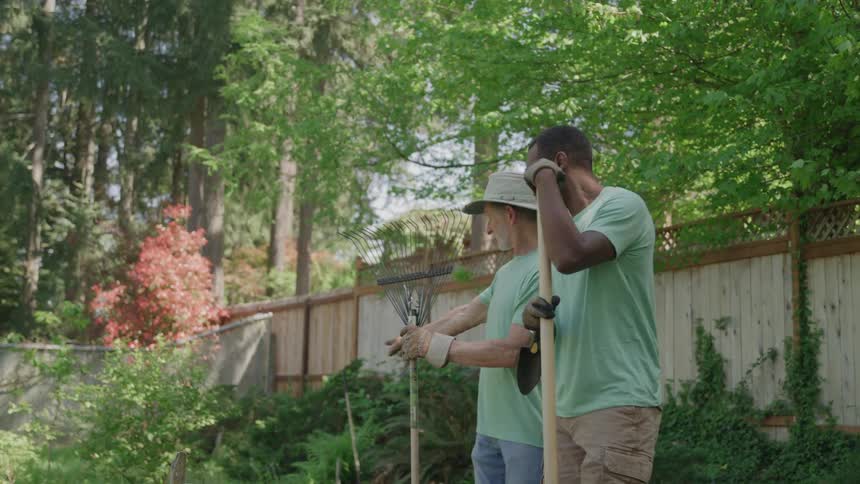 Two men wearing light green shirts and holding gardening tools stand sidebyside in a sundappled backyard with a wooden fence and dense trees forming the background