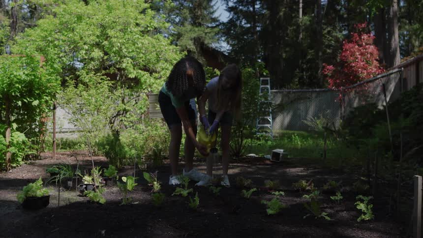 A young woman watering plants with a teenage girl in the backyard at midday