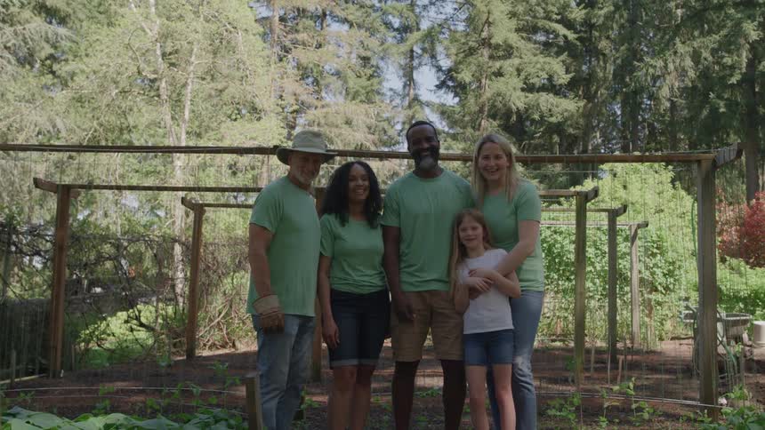 A diverse group of four adults and one young girl all wearing matching green tshirts stand together smiling at the camerathey are outdoors in a community garden or farm surrounded by growing plants and a wooden trellis structure