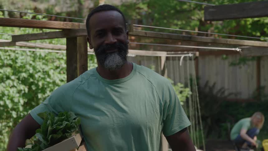 A middle age man with a gray beard wearing a light green tshirt stands in the foreground holding a wooden box overflowing with freshly harvested leafy greens framed by a rustic wooden garden structure and the blurred image of a gardener in the back