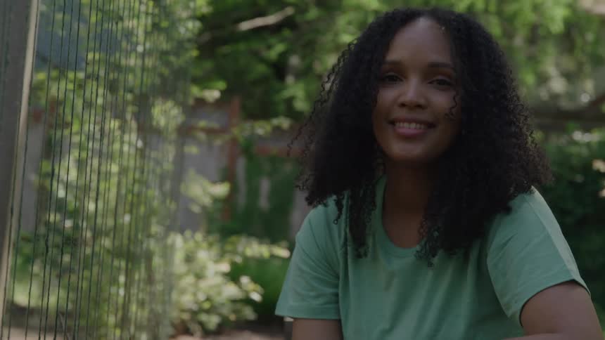 Curlyhaired woman smiles while sitting outdoors surrounded by greenery