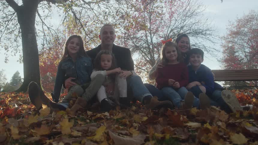 A family consisting of a man a woman and four children sitting together in a large pile of fallen autumn leaves outdoorsthey are facing the camera and smiling enjoying a sunny day in what appears to be a park with trees displaying fall colors in