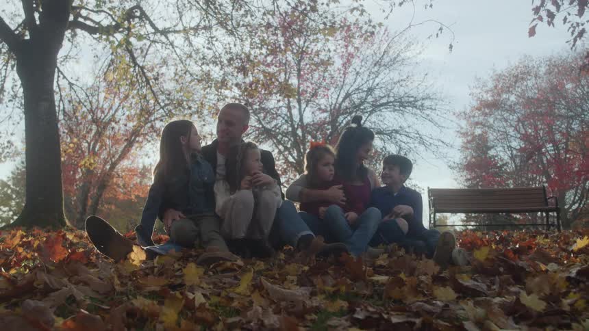 A family of six mother father and four children sits closely together on a hillside covered in a thick layer of brown and yellow fallen autumn leaves smiling and interacting in a park setting framed by trees with colorful fall foliage