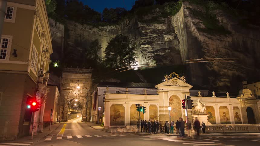 A busy city at night featuring vehicles streaking across the foreground in light trails with pedestrians gathered at a crosswalk near a baroque building and an arched tunnel entrance with a vertical rock cliff in the background