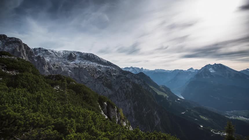 A stunning panoramic view captures a vast mountain range with rocky peaks some dusted with snow under a dramatic sky the foreground is covered with dense dark green lowgrowing vegetation overlooking a valley nestled between the slopes