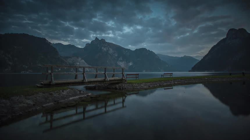 The camera is positioned low capturing a wooden pier or small bridge that extends over the water on the left with two benches visible on a small strip of land in the center the sequence transitions from late afternoon or early evening with a clear ref