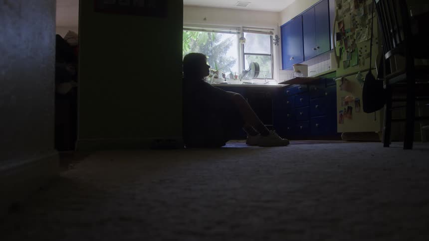 A long shot captures the silhouette of a person sitting alone on the floor of a dark room leaning against a wall with their head in their hands suggesting distress or sadness the background shows a brightly lit kitchen with blue cabinets visible throug