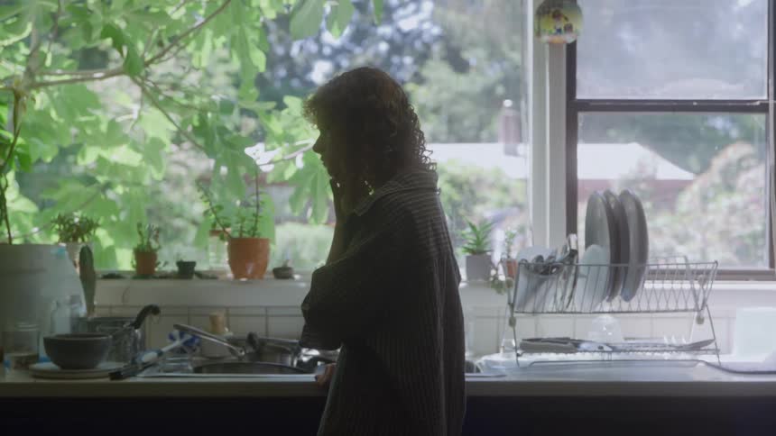 A woman stands by the kitchen sink in soft daylight surrounded by green plants