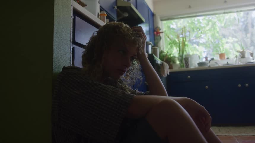 The video shows a closeup shot of a person with curly long hair sitting on the floor leaning against a dark cabinet or wall in what appears to be a kitchenthe lighting is low on the person silhouetting them against a brightly lit window that looks