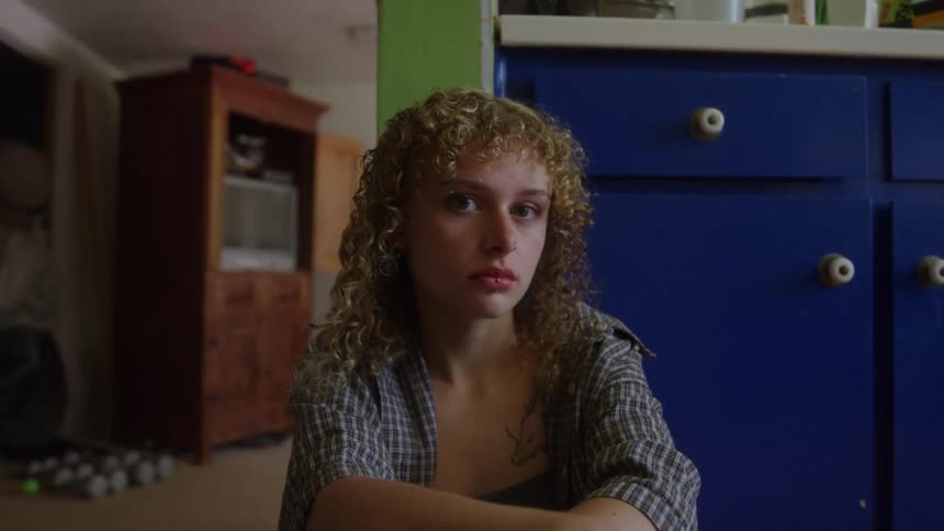 A teenage girl with curly blonde hair sitting on the floor beside a cupboard in a room at midday