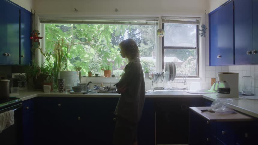A teenage girl in a white tiled kitchen nearly silhouetted against a large sunlit window that looks out onto a dense patch of green trees and yard with the person appearing contemplative