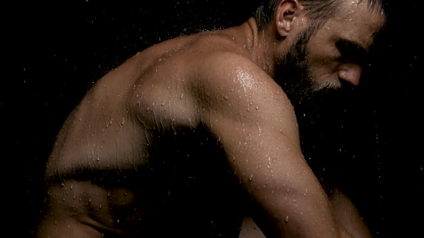 Darkly lit closeup of a shirtless man from the shoulders up facing slightly away from the camerahe appears to be standing under falling water which beads and runs down his back and shoulders with a subdued moody atmosphere