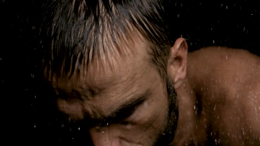 A tight closeup focuses on the face of a man with wet dark hair and a beard set against a completely black background water is visibly streaming down his face and body creating a dramatic intense look