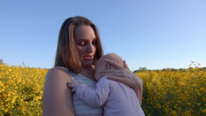 Woman holds and hugs a baby in a flower field under a blue sky