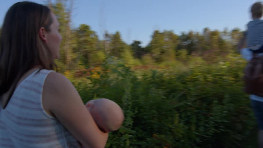 Mother carries a baby while the father carries their son on his shoulders as they walk on a path between flowering bushes on an autumn day