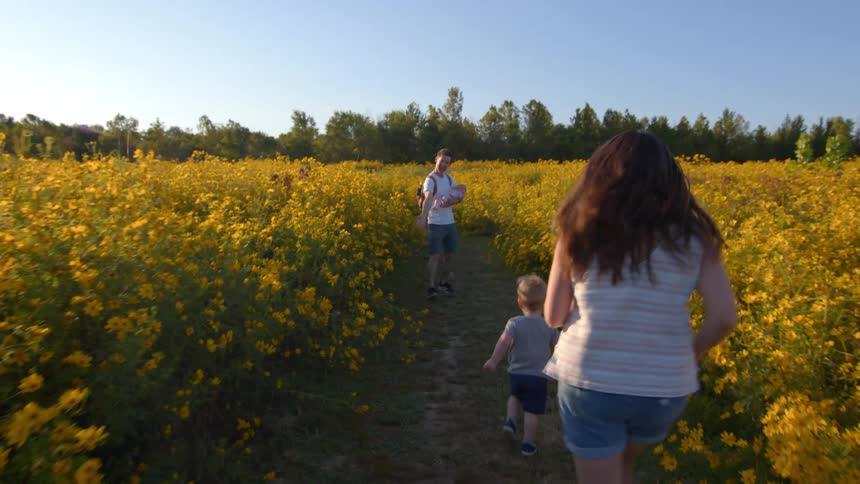 A family runs through a garden at midday with the father carrying an infant and the mother following behind a toddler