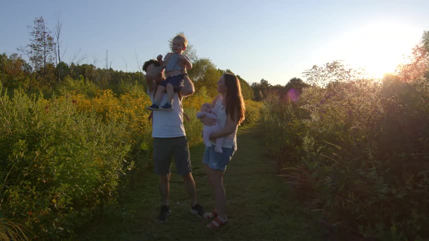 A young family consisting of a father mother and two children is standing on a dirt path in a sunny grassy field surrounded by tall weeds and yellow wildflowersthe father has one young child sitting on his shoulders while the mother holds a b