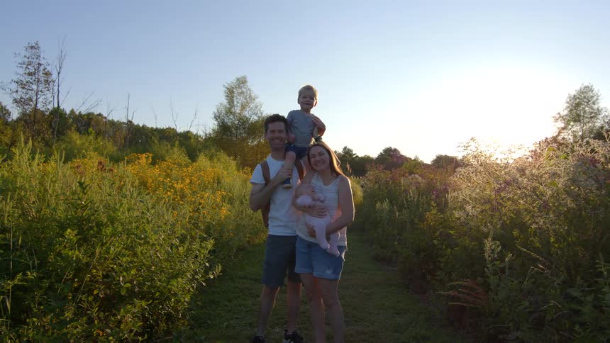 Young couple standing with their toddler male son and female baby daughter surrounded by bushes and vegetation outdoors during dusk or dawn