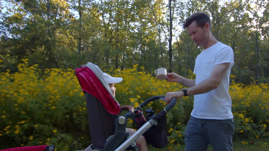 A man in a white tshirt and grey shorts is walking a baby in a red stroller through a sunny park he makes an expressive face and then takes a drink from a small cup023