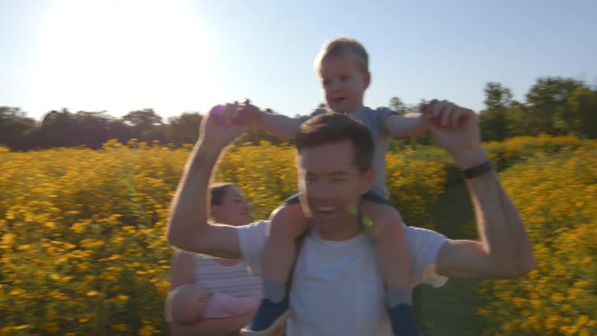 A father carries his son on his shoulders walking through an open garden field with his wife carrying an infant beside him at midday