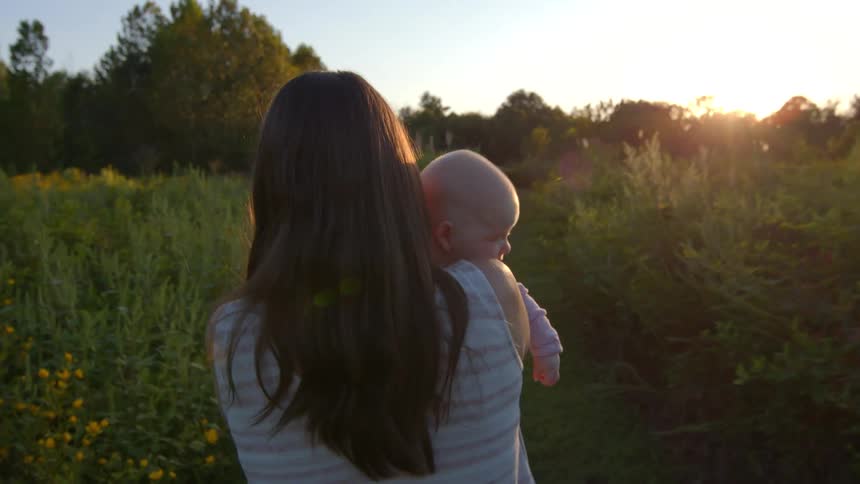 A young mother carries her baby on her shoulder while walking along a path in a farmland at sunset