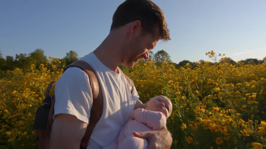 Father holds his baby in his arms in a flower field under the evening light