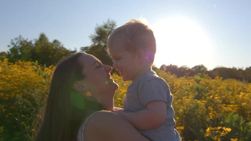 A woman is playing with her little son in her lap and they are smiling behind them is a large field with yellow flowers