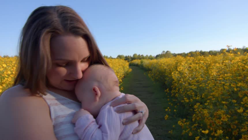 A woman with long brown hair wearing a white striped top gently holding and cuddling a baby dressed in pinkthey are standing in a vast field of bright yellow flowers with a clear blue sky overhead and a pathway visible leading through the blossoms