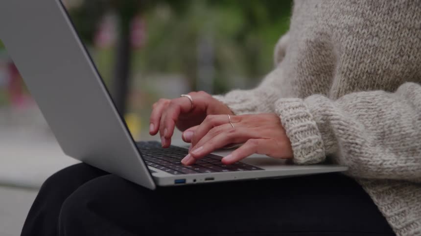 A person types on a laptop outdoors while wearing a cozy sweater