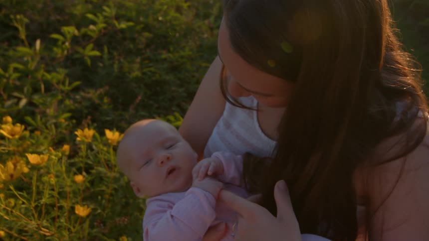 A woman with long brown hair holding and gently interacting with a baby who is dressed in a light pink outfitthe pair is outdoors bathed in warm golden light possibly during sunset and are standing near bushes with yellow flowers visible on the le