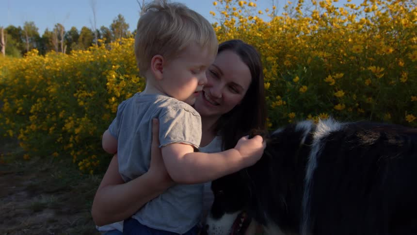 Woman holds a child as they both pet a dog in a flower field