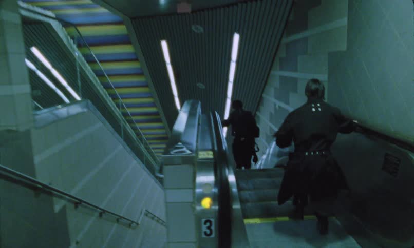 Two people wearing black run down an escalator in a transit station