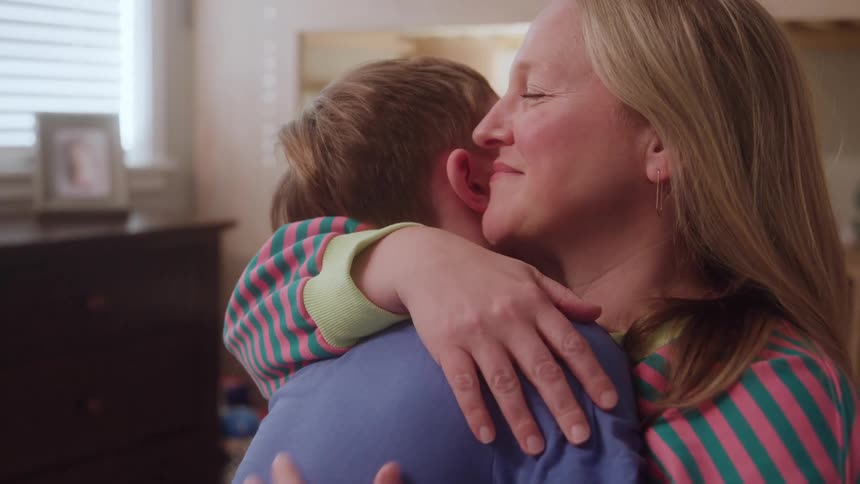 A smiling mother hugs her son warmly in a sunlit room