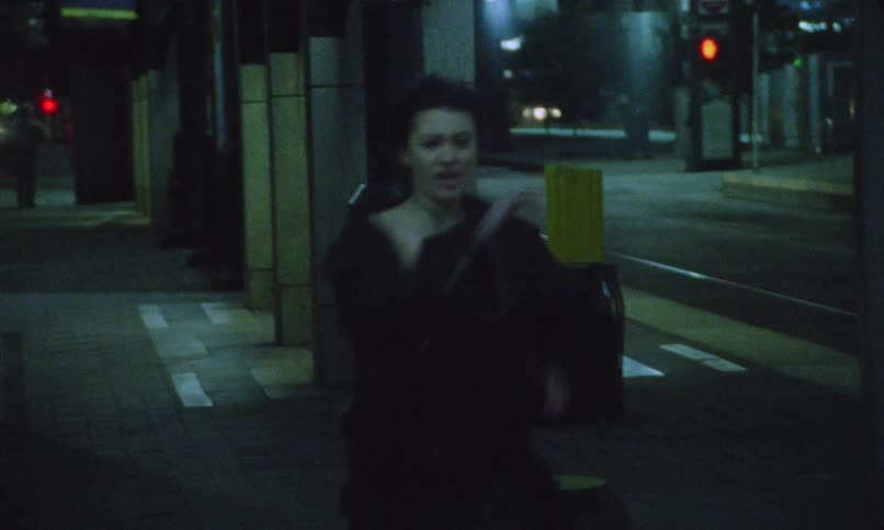 A person with dark hair dressed in dark clothing running forward through a dimly lit city street or alleyway at nightthey are running directly towards the camera with indistinct figures and street elements visible in the background suggesting a sen