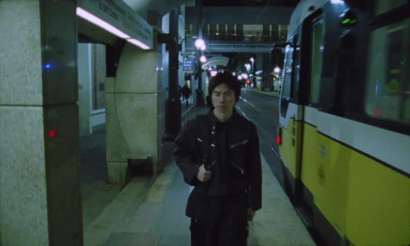 Young person with dark hair wearing a black jacket and carrying a bag over one shoulder walking along an outdoor light rail or subway platform at nightthe person walks directly toward the camera while a yellow and white train is stopped alongside the