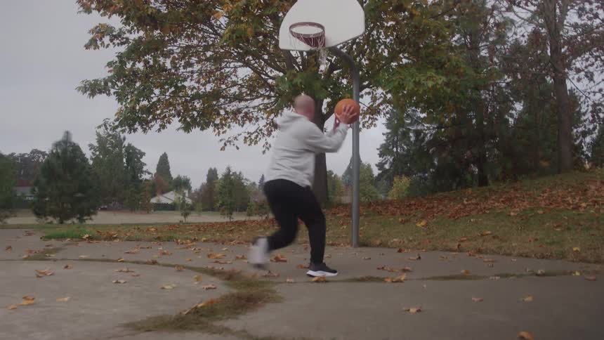 A bald man wearing a grey hoodie and black pants is playing basketball on an outdoor court covered with autumn leaves he drives toward the hoop and jumps up to successfully make a layup
