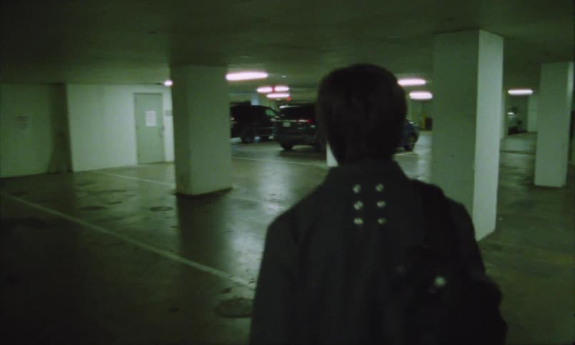 A person is standing with their back turned and wearing black clothing walking in a parking lot with several black cars in front