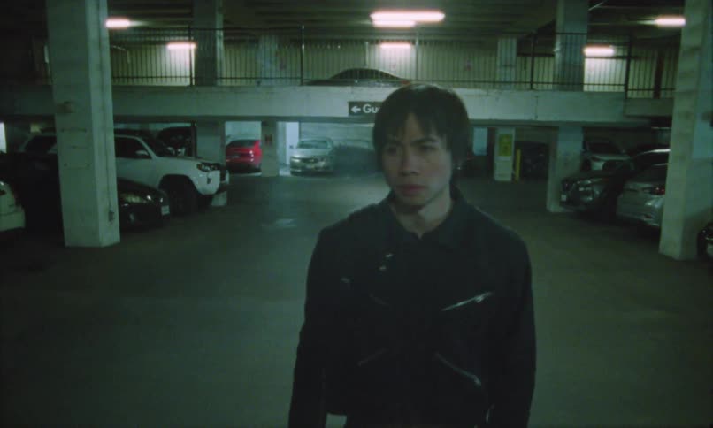 A young man wearing a jacket walks while smoking in an underground parking garage