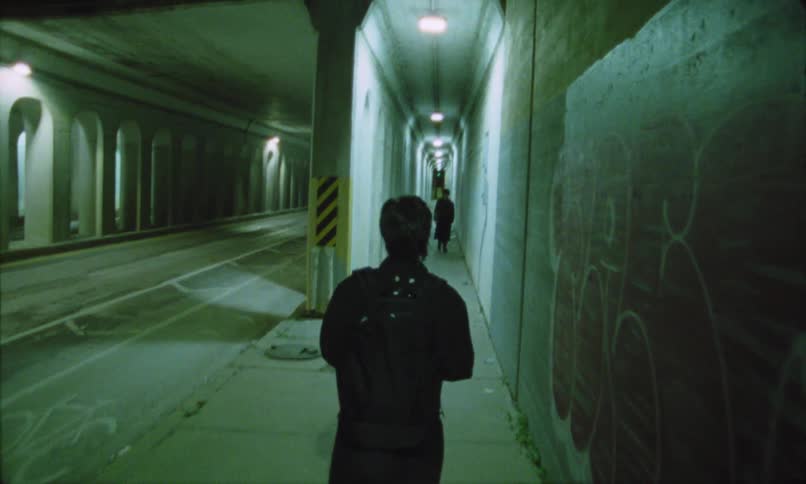 The person is wearing a dark jacket and a backpack walking towards a brightly lit far end of the passageto the left a wider parallel tunnel or road features a passing white car indicating this location is part of a larger urban infrastructure