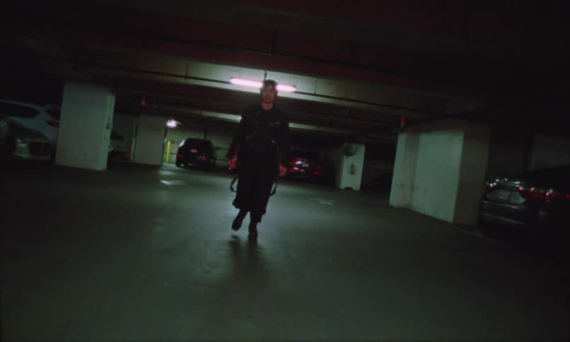 A person wearing black clothes is walking in a parking lot with several cars around