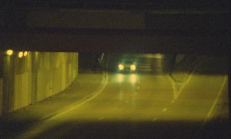 A number of cars with bright headlights speed along a wet curving road at night casting greenish reflections