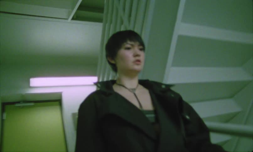 A person with short dark hair dressed in black and carrying a bag emerges from a limegreen door and begins walking up a flight of stairs the stairwell has a slightly dated institutional feel with white railings and fluorescent lighting