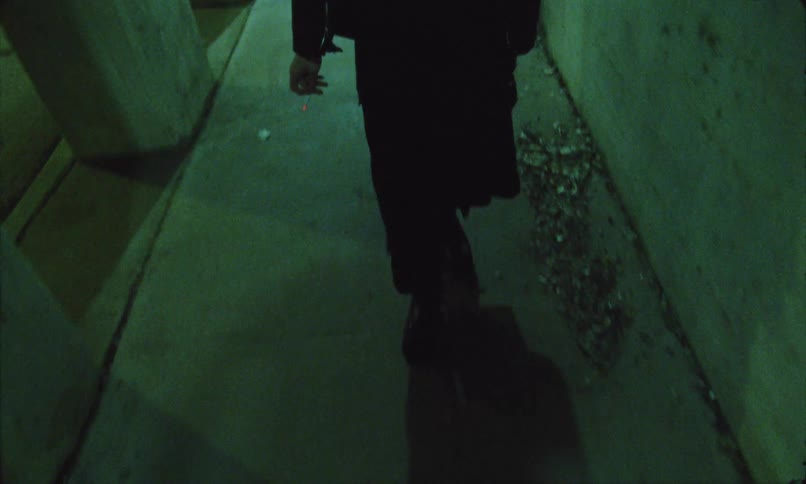 A solitary person dressed in dark clothing is captured from a low angle as he walk away down a narrow concrete urban passageway dimly lit with an eerie green hue casting a long distinct shadow ahead of his feet
